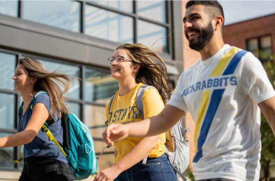 Three students, two females and a male, wearing SD State shirts, walk smiling through a courtyard. The two women have brown hair that is blowing in the wind and the male has short and cropped hair and a beard.