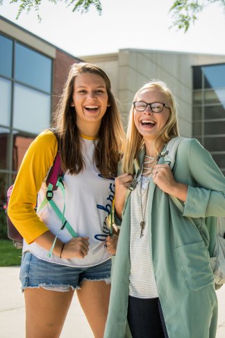Two white women laughing and smiling towards the camera. One is brunette and wears a yellow and white t-shirt and shorts, the other is blond with glasses and greenish-blue sweater