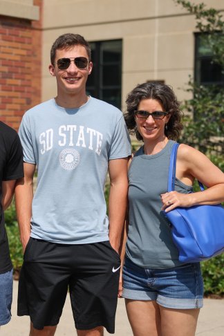 A white male student wearing a light blue "SD State" t-shirt and shorts and sunglasses, and his mother, wearing a gray shirt and jean shorts carrying a blue bag. Both are smiling for the camera.