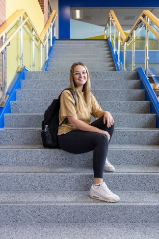 Photo of a smiling blond, white female student sitting on steps, wearing a yellow shirt, black pants and white sneakers. The stair rails are yellow and blue.
