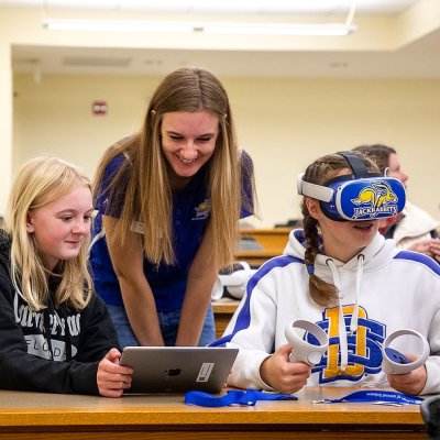 An image of a student using a VR headset and 2 other students monitoring a tablet connected to the display