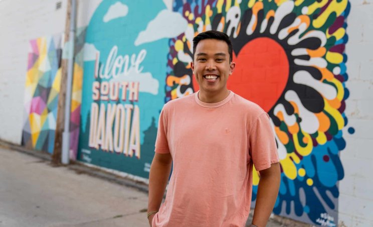 A smiling man in a pink shirt stands in front of a mural wall that says "I love South Dakota"