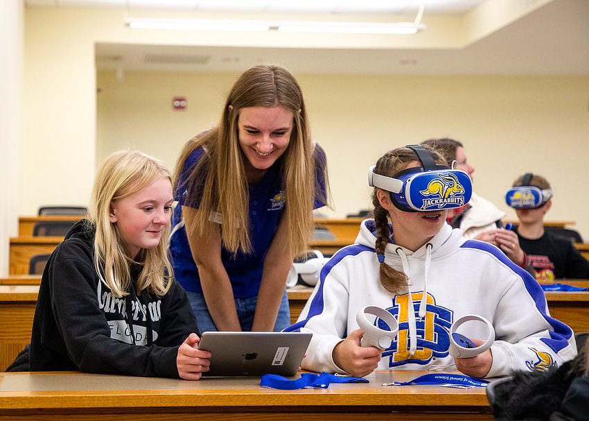 An image of a student using a VR headset and 2 other students monitoring a tablet connected to the display