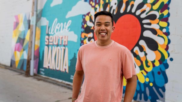 A smiling man in a pink shirt stands in front of a mural wall that says "I love South Dakota"