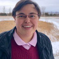 Picture of a person with short black hair, glasses, in a pink shirt and maroon sweater in front of prairie grass and snow